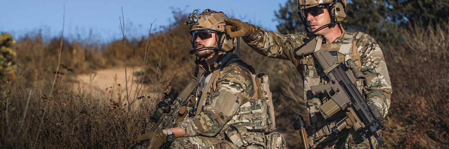 Two soldiers in camouflage uniforms and tactical gear, one kneeling and the other standing while pointing, in a dry, grassy outdoor environment.