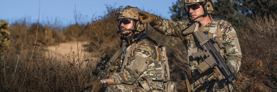 Two soldiers in camouflage uniforms and tactical gear, one kneeling and the other standing while pointing, in a dry, grassy outdoor environment.