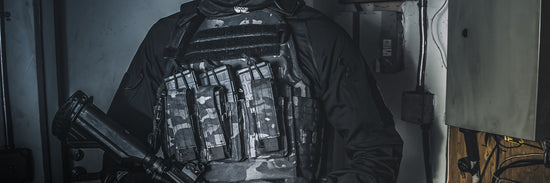 Person in tactical gear holding a firearm, wearing a camouflage vest with multiple magazine pouches, standing indoors near electrical panels.