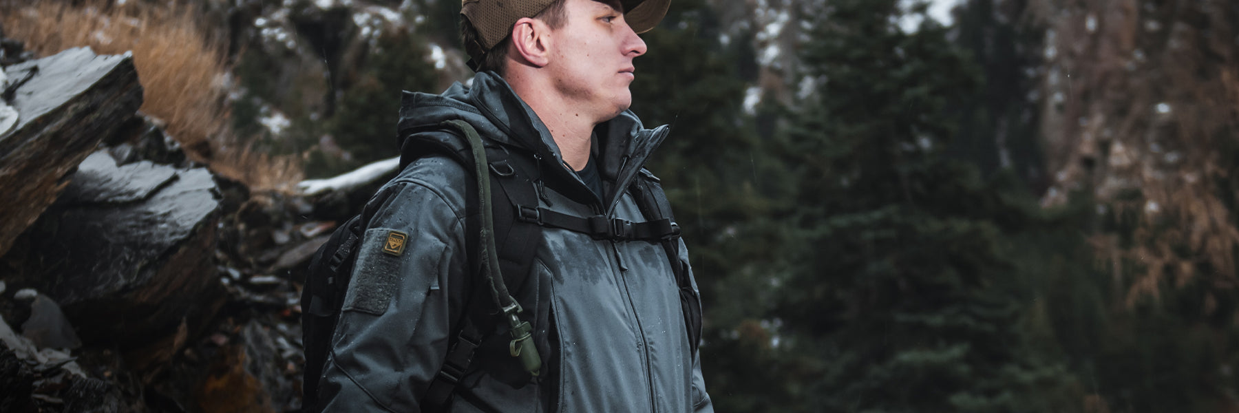 A man in outdoor gear and a cap stands on a rocky path with evergreen trees and snow-dusted mountains in the background.