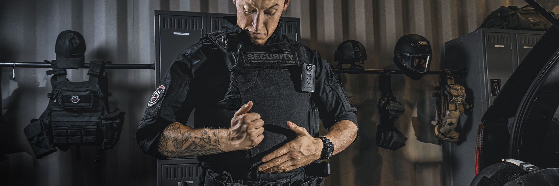 A security officer in tactical gear adjusts their vest in a locker room, with helmets and equipment visible in the background.