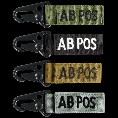 The Condor Blood Type Key Chain set includes four keychains embroidered with “AB POS” in green, black, tan, and grey, each featuring a black metal clip.