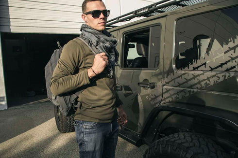 A man in the Condor Base II Crew Pullover—ideal for EDC fans—with sunglasses, scarf, and backpack stands by a green geometric-patterned off-road vehicle outside a building.