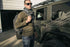 A man in the Condor Base II Crew Pullover—ideal for EDC fans—with sunglasses, scarf, and backpack stands by a green geometric-patterned off-road vehicle outside a building.