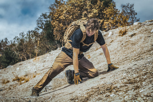 Wearing Condors Paladin Tactical Pants, a person with a backpack kneels on rocky, sloped terrain with trees and a cloudy sky in the background.