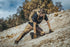 Wearing Condors Paladin Tactical Pants, a person with a backpack kneels on rocky, sloped terrain with trees and a cloudy sky in the background.