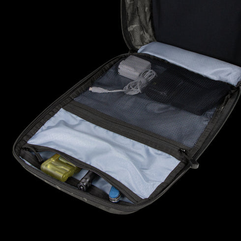 The Condor Prime Pack 21L - MultiCam Black is open on a white surface, revealing mesh pockets with a power adapter, cables, and small tools; wooden blocks are visible in the background.
