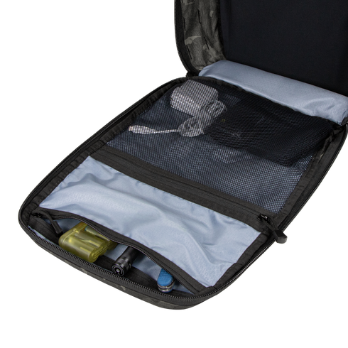 The Condor Prime Pack 21L - MultiCam Black is open on a white surface, revealing mesh pockets with a power adapter, cables, and small tools; wooden blocks are visible in the background.