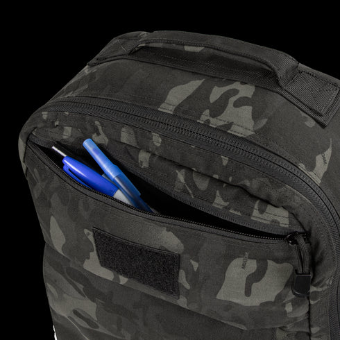 A close-up of the Condor Prime Pack 21L in MultiCam Black with its front zipper pocket open, revealing two blue pens and one black pen inside.