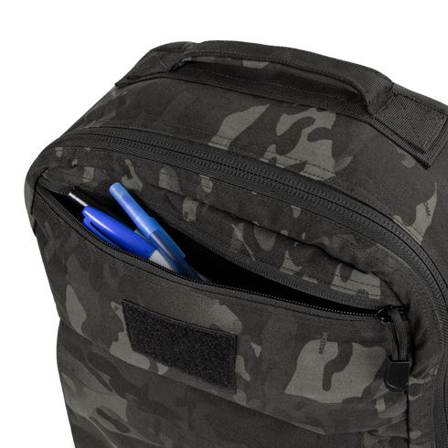 A close-up of the Condor Prime Pack 21L in MultiCam Black with its front zipper pocket open, revealing two blue pens and one black pen inside.