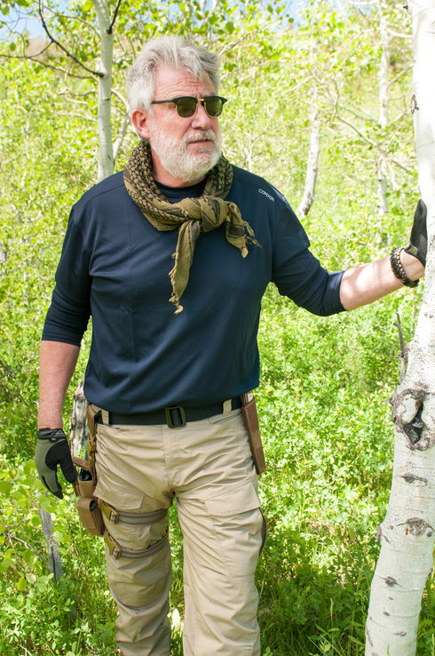 A man with gray hair and a beard, wearing the Condor Maxfort Long Sleeve Training Top, tan cargo pants, and gloves, stands outdoors in a green wooded area holding a tree, demonstrating his bushcraft skills.