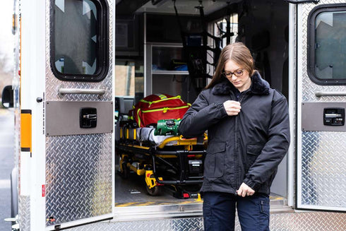 A person stands outside an open ambulance, zipping up a Condor Guardian Duty Jacket | SALE. The ambulance interior and a stretcher with medical supplies are visible behind them.