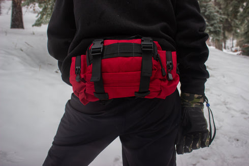 A person stands in a snowy forest wearing black gear and gloves, with a large red Condor Deployment Bag 6.5L strapped at the waist—ideal for first responders or law enforcement professionals on duty.