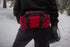 A person stands in a snowy forest wearing black gear and gloves, with a large red Condor Deployment Bag 6.5L strapped at the waist—ideal for first responders or law enforcement professionals on duty.