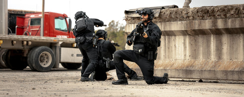 Three armed tactical police officers in Condor Paladin Tactical Pants take cover behind a concrete barrier and a truck during a daytime operation.