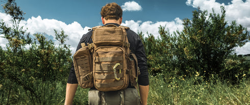 A person with a large tan Condor Field Pouch stands outdoors facing dense green vegetation beneath a partly cloudy sky.