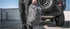 A person in dark pants holds a Condor Elite Fail Safe Urban Backpack Gen II 18L by Condor, standing next to the rear of an off-road vehicle with a spare tire.