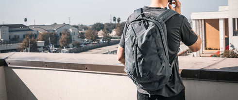 A person wearing the Condor Elite Fail Safe Urban Backpack Gen II 18L by Condor stands on a rooftop terrace, facing away and talking on a phone, with buildings and trees in the background.