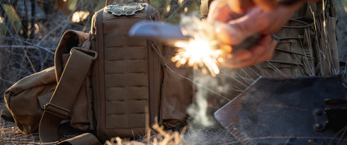 A person strikes a fire starter, creating sparks beside a Condor Field Pouch and a shovel on dry grass outdoors.