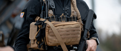 Close-up of a person in tactical gear, featuring a Condor Single M4 Mag Pouch on their tan chest rig, holding magazines and a radio, with gloves and an American flag patch on their sleeve.