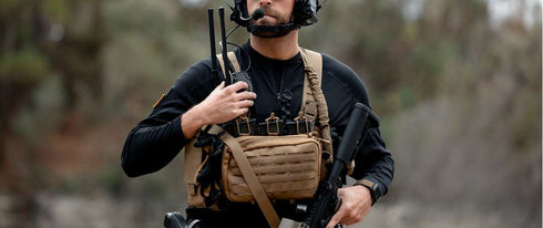 A person in tactical gear, wearing a Condor Single M4 Mag Pouch, headset, vest, and holding a rifle stands outdoors with a radio device.