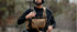 A person in tactical gear, wearing a Condor Single M4 Mag Pouch, headset, vest, and holding a rifle stands outdoors with a radio device.
