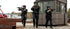 Three armed tactical police officers in Condor Paladin Tactical Pants and black gear advance with weapons raised outside a guard booth in an industrial area.