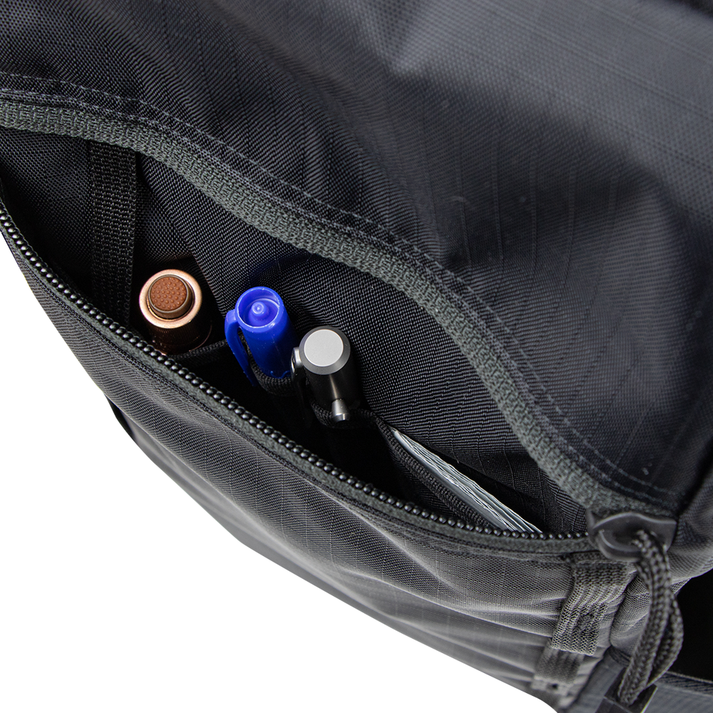 Close-up of the open zippered pocket on the Condor Aero Roll-Top Backpack 25L, holding a brown pen, blue pen, black pen, and metallic gray pen—gear favored by military and first responders.