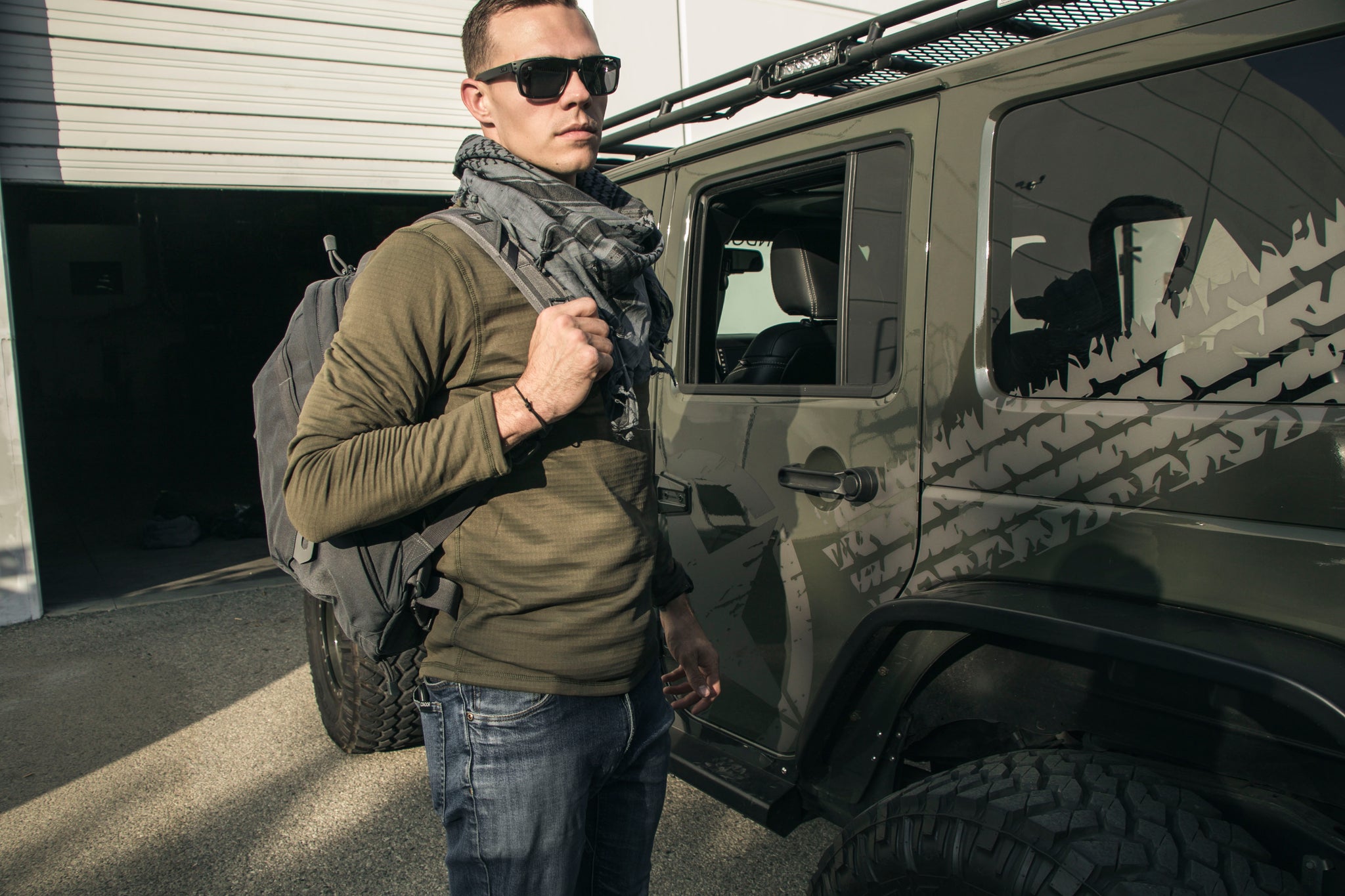 A man in the Condor Base II Crew Pullover—ideal for EDC fans—with sunglasses, scarf, and backpack stands by a green geometric-patterned off-road vehicle outside a building.