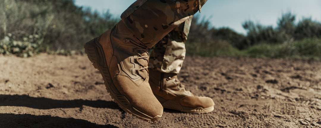 A person wearing Condor Brigade 8″ Tactical Boots – AR670 and camouflage pants stands on a dirt path outdoors, surrounded by vegetation and under a blue sky.