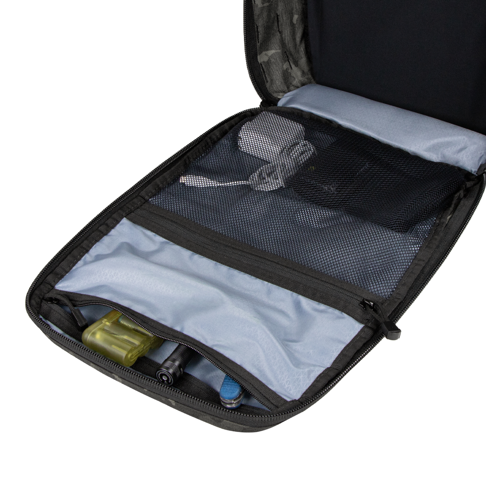 The Condor Prime Pack 21L - MultiCam Black is open on a white surface, revealing mesh pockets with a power adapter, cables, and small tools; wooden blocks are visible in the background.