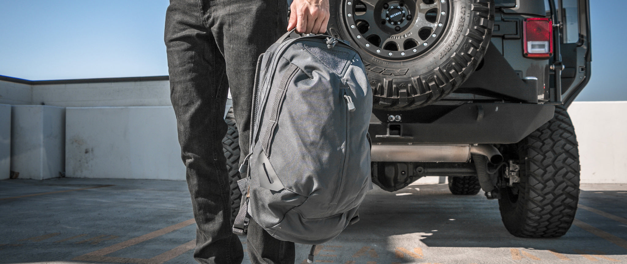 A person in dark pants holds a Condor Elite Fail Safe Urban Backpack Gen II 18L by Condor, standing next to the rear of an off-road vehicle with a spare tire.