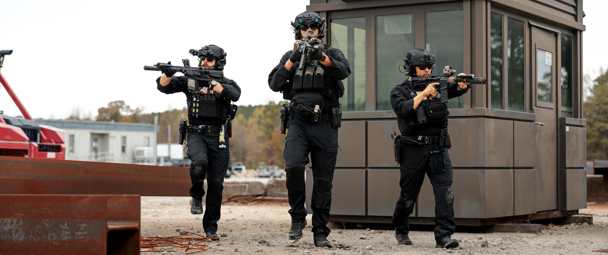 Three armed tactical police officers in Condor Paladin Tactical Pants and black gear advance with weapons raised outside a guard booth in an industrial area.